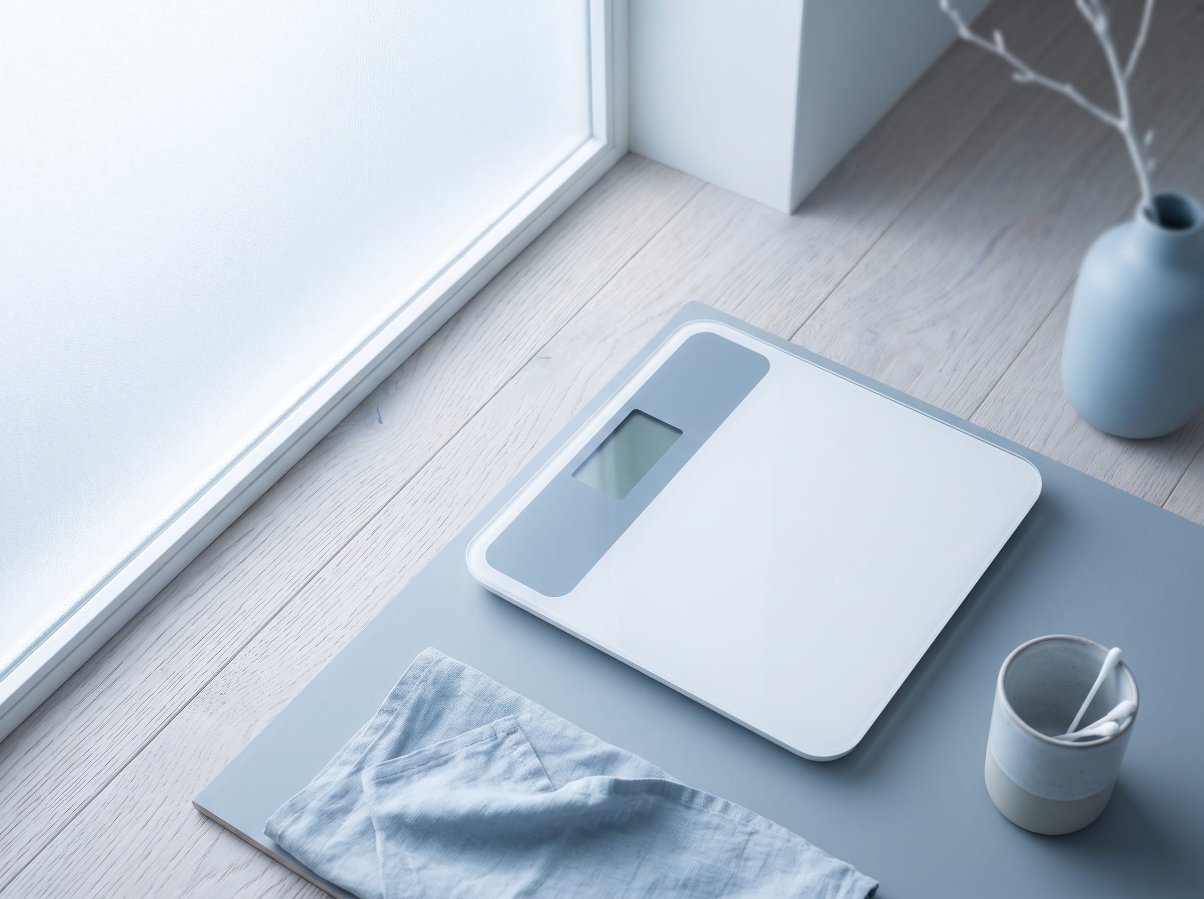 A digital scale on a pale wood floor beside a window, seen from above